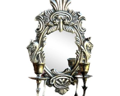 Rustic European mirrored wall sconce with two candleholders. The central frame features a large oval opening surrounded by rococo floral motifs, including leaves, berries, and shell-like designs. Two candle holders extend from the center, each supported by curved arms and finished with small drip trays for candles.. Dimensions 7.5 W 3.5 D 11 H Styles Baroque Rococo Rustic European Period 1970s ...