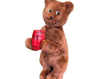 Charming mid-20th century plush bear toy, featuring soft brown fur and inset plastic eyes.The bear holds a painted red wooden barrel with a hand-painted inscription. The back shows where a small winding mechanism should be, suggesting this was once a mechanical or windup toy, likely designed to create simple movements when activated. The bear stands upright with a slightly whimsical expression,...