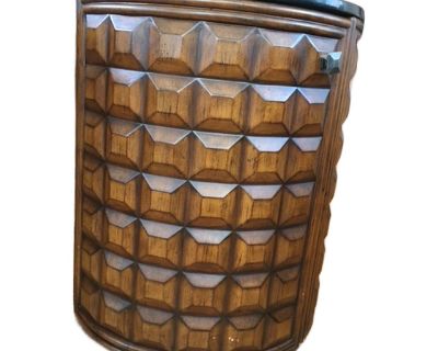 Sculptural Mid-Century Modern drum-form dry bar featuring a bold, geometric carved walnut exterior and polished black stone top. The deep, faceted carving evokes Brutalist design influences of the 1970s, giving this piece strong presence as both furniture and art. A concealed, hinged door opens to reveal curved interior shelving, stemware racks, and dedicated bottle storage. The bar rests on sm...